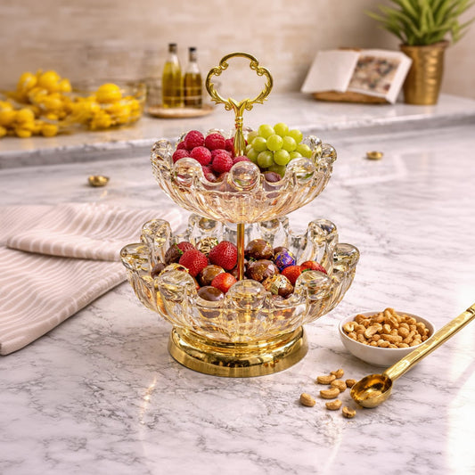 Two-tiered glass fruit bowl with gold base filled with fruits and nuts on a marble surface.