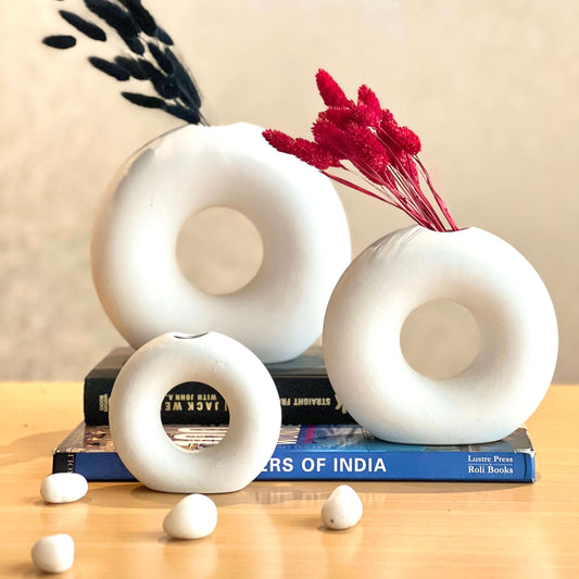 White donut-shaped vases on books with red dried flowers and small white stones on a wooden surface.